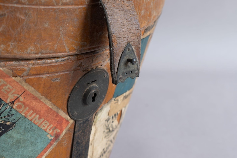 May include: Close-up of a vintage brown leather travel trunk with a metal lock and handle. The trunk has several colorful travel stickers, including one that says "NEW COLUMBUS". The trunk is weathered and shows signs of age.
