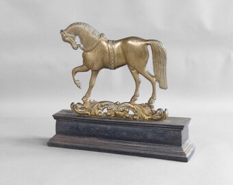 May include: A brass horse sculpture with intricate detailing, standing on a dark, rectangular base. The horse is posed with one leg raised, and the base features ornate, swirling patterns. The sculpture is a decorative item.