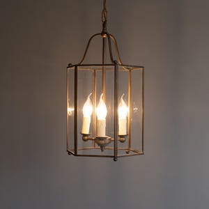 K&ouml;nnte beinhalten: Eine sechseckige Laternen-H&auml;ngeleuchte mit einem dunkelbronzenen Metallrahmen und klaren Glasscheiben. Die Leuchte verf&uuml;gt &uuml;ber drei interne Leuchtmittel im Kerzenstil mit wei&szlig;en Abdeckungen, die an einer passenden Metallkette h&auml;ngen.