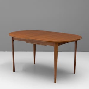 Puede incluir: Una mesa de comedor ovalada fabricada en madera de tono medio con una superficie lisa y pulida. La mesa cuenta con cuatro patas cónicas que se extienden hacia afuera desde el centro, proporcionando una base estable para el tablero.