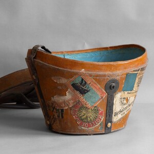 May include: A vintage, brown leather bucket-shaped container with a blue interior. The exterior is adorned with travel stickers and a metal clasp. A matching lid rests nearby. The container has a handle.