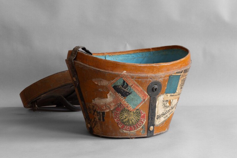 May include: A vintage, brown leather bucket-shaped container with a blue interior. The exterior is adorned with travel stickers and a metal clasp. A matching lid rests nearby. The container has a handle.