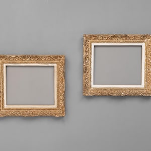 May include: Two antique-style picture frames with gold-coloured, detailed borders and square openings. The frames have a white inner edge. The background is a plain grey colour.