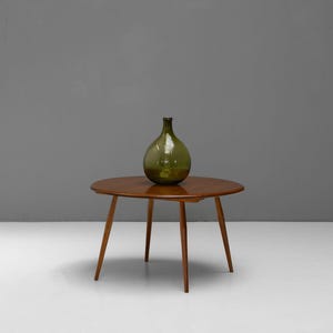 May include: A round, wooden coffee table with three angled legs. A large, green glass bottle sits on the table's surface. The table and bottle are set against a neutral gray background.