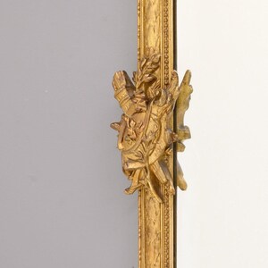 May include: A gold-colored ornate mirror frame with a detailed carved design. The frame features a sculpted design of a figure holding a wreath and a sword.