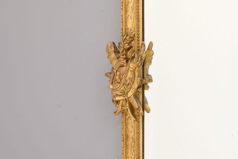 May include: A gold-colored ornate mirror frame with a detailed carved design. The frame features a sculpted design of a figure holding a wreath and a sword.