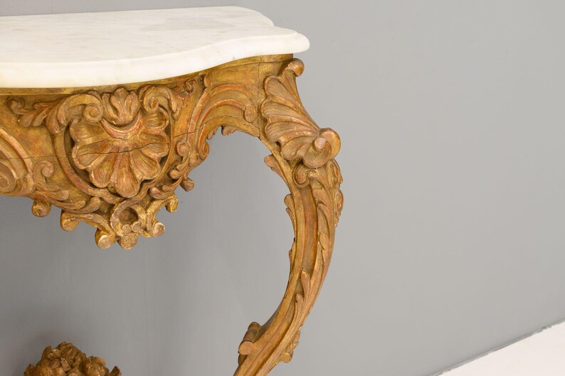 Peut inclure: Une console en bois peinte en or avec un dessus en marbre blanc et des sculptures complexes. La table a une jambe incurv&eacute;e et orn&eacute;e.