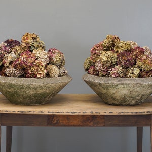 Op de afbeelding: Twee grote, verweerde, grijze betonnen plantenbakken met een brede, ondiepe komvorm. Elke plantenbak is gevuld met een groot boeket gedroogde hortensia's in tinten roze, crème en beige. De plantenbakken staan op een rustieke houten tafel.