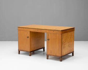 May include: A light brown wooden desk with two cabinets and a central open space. The desk has a rectangular top and is supported by short, dark-colored legs. The cabinets feature small, round handles.