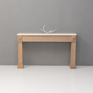May include: A light-coloured wooden console table with a white marble top. The table has four square legs and decorative details on the front. A clear, abstract glass sculpture sits on top of the table.