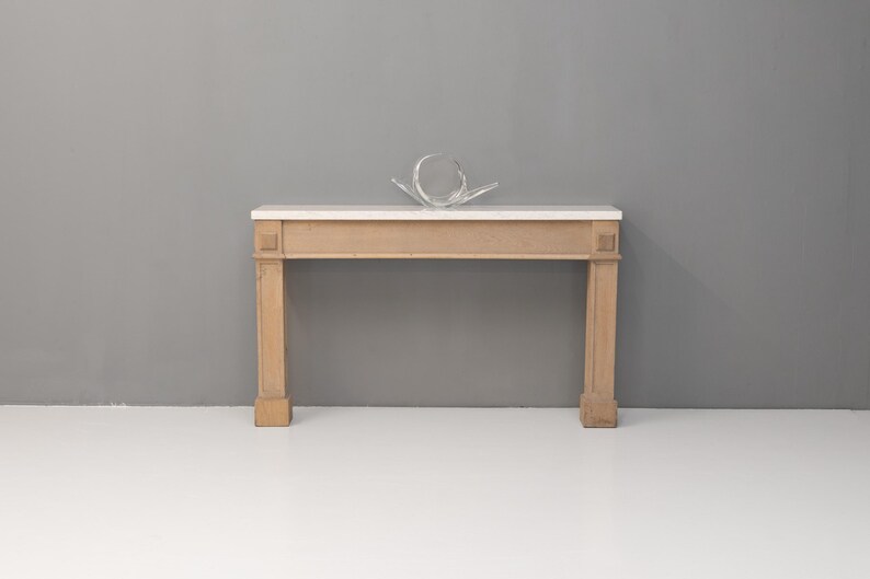 May include: A light-coloured wooden console table with a white marble top. The table has four square legs and decorative details on the front. A clear, abstract glass sculpture sits on top of the table.