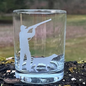 Puede incluir: Un vaso de vidrio transparente con un diseño grabado de un cazador con un arma y un perro. El diseño es blanco y el vidrio es transparente.