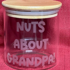 Peut inclure: Un pot en verre transparent avec un couvercle en bois. Le pot porte l'inscription "NUTS ABOUT GRANDPA!" en rose.