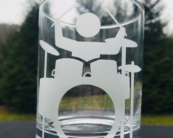 May include: A clear glass tumbler with a white etched design of a drummer playing a drum set. The design is simple and minimalist, with the drummer's figure and the drums outlined in white.