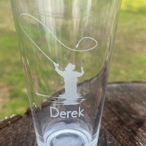 May include: A clear glass with an etched design of a person fly fishing. The glass has the name "Derek" etched at the bottom.