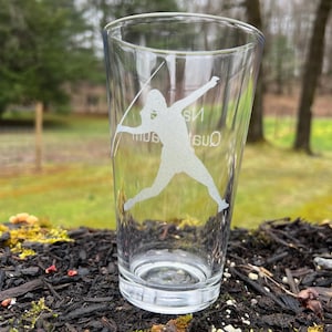 Op de afbeelding: Een helder glazen pintglas met een wit silhouet van een persoon die een speer werpt, gegraveerd aan de zijkant. De tekst "Quata" is gegraveerd op het glas.