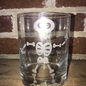 May include: A clear glass tumbler featuring a white etched design of a skeleton. The skeleton is depicted in a playful pose with its arms outstretched and legs crossed.