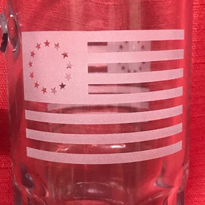 Könnte beinhalten: Ein klares Glasbierkrug mit einem geätzten Design der Betsy Ross Flagge. Die Flagge hat 13 Sterne in einem Kreis und 13 rote und weiße Streifen.