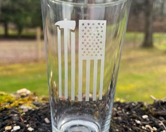 Puede incluir: Un vaso de pinta de vidrio transparente con un diseño grabado de una bandera estadounidense y un hacha. El vaso está sentado en un lecho de mantillo marrón.