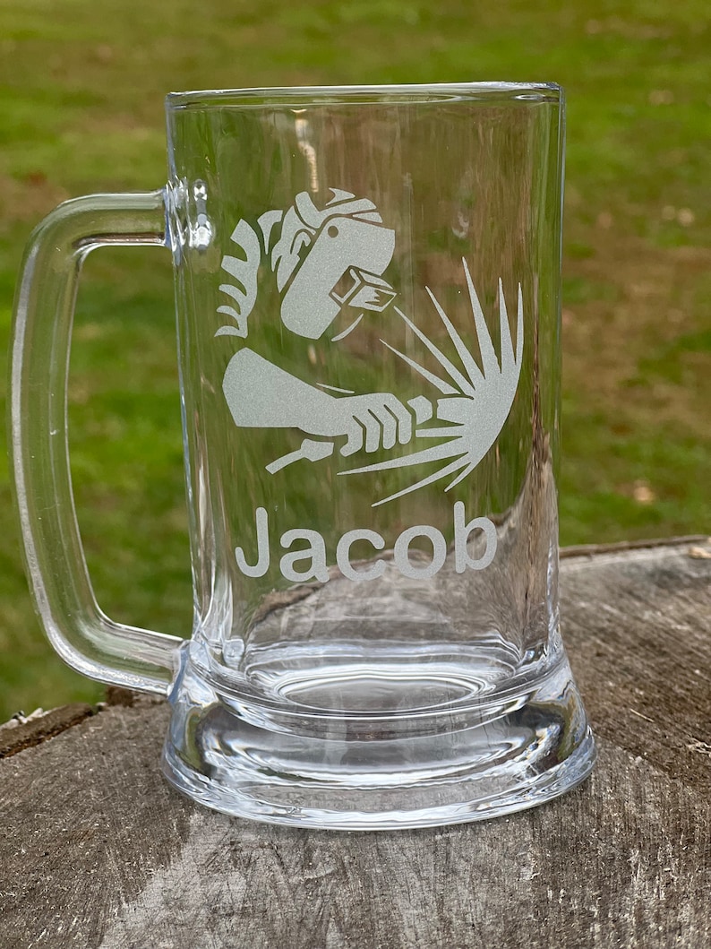 May include: A clear glass beer stein with a white etched design of a welder and the name "Jacob".