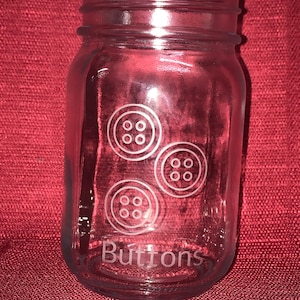 May include: A clear glass jar with a wide mouth and a metal lid. The jar is etched with three buttons and the word "Buttons".