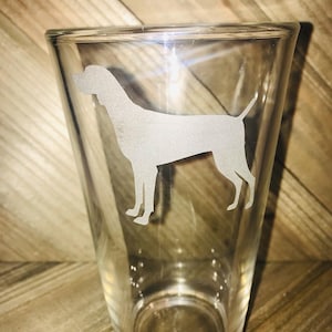May include: A clear glass pint glass with an etched silhouette of a dog. The dog is facing the right side of the glass.
