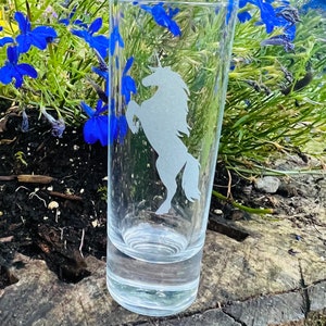 Puede incluir: Un vaso de vidrio transparente con un unicornio grabado en el lateral. El vaso está sobre una superficie de madera.