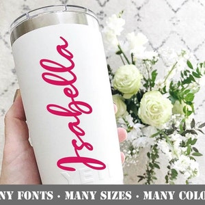 May include: A white tumbler with the name "Isabella" in a vibrant pink script. The text "MANY FONTS • MANY SIZES • MANY COLORS" is at the bottom. The tumbler is held in a hand, with a bouquet of white flowers in the background.