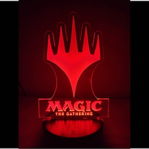 May include: A red LED night light shaped like the Magic: The Gathering symbol. The symbol is a five-pointed star with a diamond in the center. The words "Magic: The Gathering" are printed below the symbol.