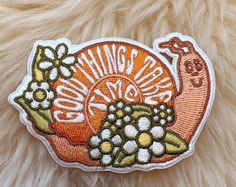 May include: A white embroidered patch with a snail design. The snail is orange and has the words "Good Things Take Time" written on its shell. The snail is surrounded by white flowers.