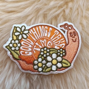 May include: A white embroidered patch featuring a snail design. The snail is orange and has the words "Good Things Take Time" written on its shell. The snail is surrounded by white flowers.