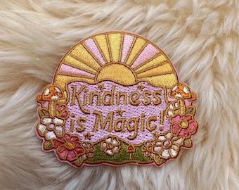 May include: A pink and gold embroidered patch with a sun and the text "Kindness is Magic!" surrounded by flowers and mushrooms.