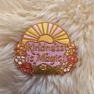 May include: A pink and gold embroidered patch with a sun and the text "Kindness is Magic!" surrounded by flowers and mushrooms.