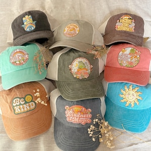 Puede incluir: Nueve gorras de camionero diferentes con varios diseños bordados. Los diseños incluyen una mano con un ojo, una rana, un sol, una cara sonriente y texto que dice "No te preocupes", "Disfruta del viaje", "La amabilidad es mágica", "Sé amable" y "Exactamente donde necesitas estar".
