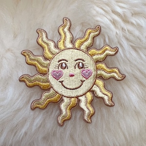 May include: A yellow embroidered sun with a smiling face and pink hearts for cheeks. The sun has a detailed design with many rays.