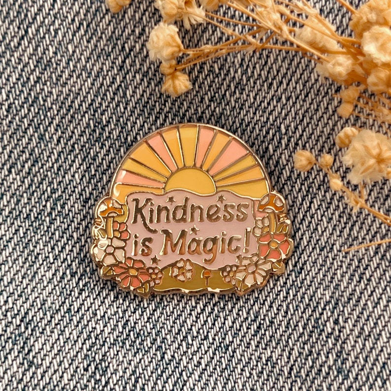 May include: A gold enamel pin featuring a pink and yellow sunset design with the text "Kindness is Magic!" surrounded by flowers and mushrooms.