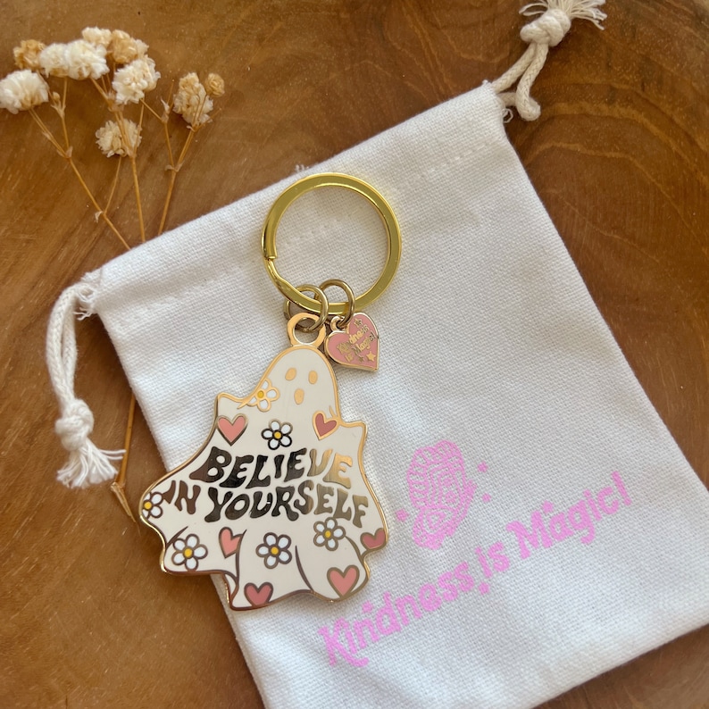 Puede incluir: Un llavero dorado con un dije en forma de fantasma que dice "Believe in Yourself" en letras doradas. El dije es blanco con corazones y flores rosas. Un peque&ntilde;o dije de coraz&oacute;n rosa cuelga debajo. El llavero descansa sobre una bolsa blanca.