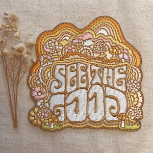 May include: Embroidered patch with the words "SEE THE GOOD" in a retro font. The patch features a wavy, floral design in shades of gold, orange, pink, and white. The patch is likely for clothing or accessories.