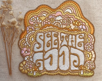 May include: Embroidered patch with the words "SEE THE GOOD" in a retro font. The patch features a wavy, floral design in shades of gold, orange, pink, and white. The patch is likely for clothing or accessories.