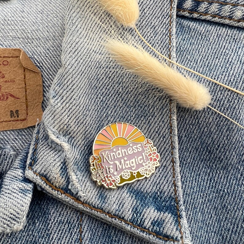 May include: A colourful enamel pin with a sun and flowers surrounding the text "Kindness is Magic!"