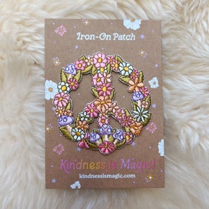 May include: Iron-on patch featuring a peace sign made of embroidered flowers in shades of pink, white, and yellow. The patch is on a brown card with the text "Iron-On Patch" and "Kindness is Magic!" with the website address "kindnessismagic.com".
