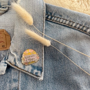 May include: A gold enamel pin with a pink and yellow sun and flowers. The pin says "Kindness is Magic".