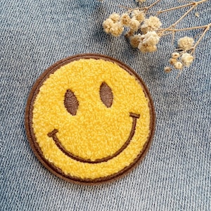 May include: A yellow smiley face patch with brown stitching, made of fuzzy fabric. The patch is on a blue denim background.