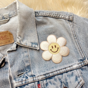 Puede incluir: Una chaqueta vaquera azul claro con un parche bordado de flor blanca con una carita sonriente amarilla.
