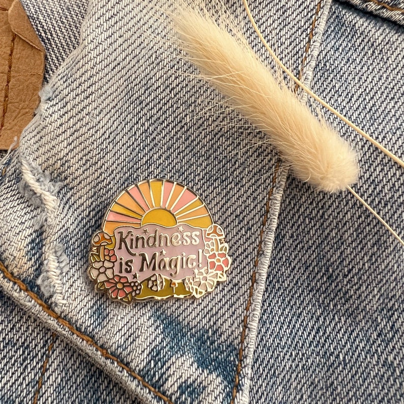 May include: A gold enamel pin featuring a pink and yellow sunset design with the text "Kindness is Magic!" surrounded by flowers and mushrooms.