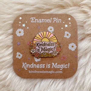 May include: A gold enamel pin featuring a pink and yellow sunset design. The pin reads "Kindness is Magic!" and has a floral border. The pin is displayed on a brown card with the same text and the website address "kindnessismagic.com".