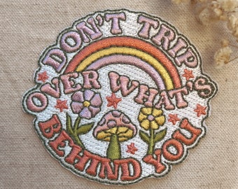 May include: A white embroidered patch with a rainbow, flowers, and mushrooms. The text reads "Don't Trip Over What's Behind You".