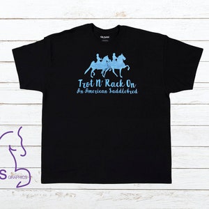 Op de afbeelding: Zwart T-shirt met een blauwe afbeelding van drie paarden en ruiters. De tekst "Trot N' Rack On An American Saddlebred" is onder de afbeelding gedrukt.