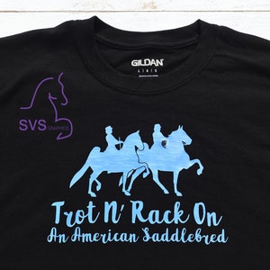 Op de afbeelding: Zwart T-shirt met een lichtblauwe afbeelding van drie paarden en ruiters. De tekst "Trot N' Rack On An American Saddlebred" is onder de afbeelding gedrukt.
