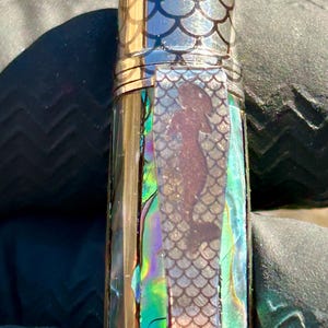 Puede incluir: Primer plano de una pluma con un diseño de sirena. La pluma está hecha de concha de abulón iridiscente y tiene una banda dorada alrededor de la parte superior. El diseño de la sirena está en una silueta oscura contra el fondo iridiscente.